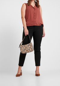 Blouse sans manches en rouille avec un col en V et des boutons, associée à un pantalon slim noir. Accessoire comprenant un sac à main en imprimé serpent beige et marron.