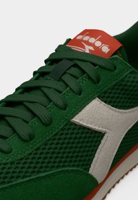 Κοντινό πλάνο πράσινου αθλητικού παπουτσιού με λευκό λογότυπο στο πλάι, σκούρους πράσινους κορδόνια και πορτοκαλί ετικέτα στη γλώσσα με τη μάρκα «diadora».