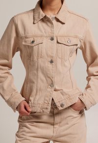 Ljusbeige jeansjacka med klassisk krage, två bröstfickor, knappar framtill och en figursydd design. Har synliga sömmar och en slät textur.