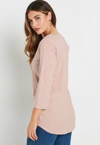 Haut à manches longues rose pâle avec un col rond, texture douce, fentes latérales et ourlet arrondi. Associé à un jean bleu foncé.