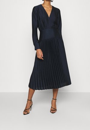 Vrouw draagt een zwarte, geplooide midi-jurk met lange mouwen en diepe V-hals, en zwarte enkellaarsjes met hoge hak en enkelbandje, staande op een grijze vloer.