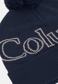 Jūras spēku adīta beanie cepure ar mīkstu pom-pom augšpusē. Iekļauj kontrastējošas gaiši pelēkas burtus gar priekšpusi. Ribiņu apkakle apakšā.