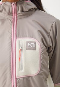 Απαλό ροζ και καφέ μπουφάν με θήκη από φλις και αντίθετες ροζ λεπτομέρειες. Διαθέτει ψηλό γιακά και φερμουάρ με πορτοκαλί λαβή.