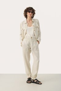 Linen outfit in off-white, featuring a button-up jacket, loose trousers with pockets, and black strappy sandals. Includes round sunglasses.