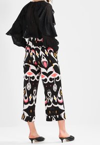 Femme portant un chemisier noir transparent et un pantalon large court à motifs noirs, blancs et roses, avec des mules noires à talons, debout les mains dans les poches.