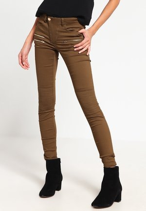 Kvinde iført brune smalle bukser med lynlåslommer foran og ribstrikkede knædetaljer, kombineret med sorte ankelstøvler og en sort top.