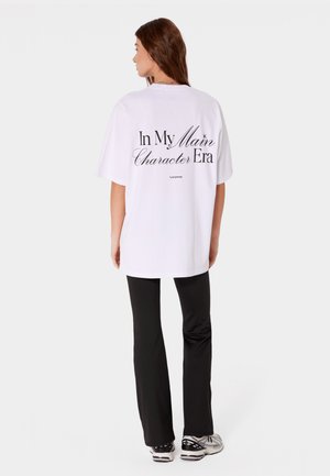 Kobieta w białym, oversize T-shircie z napisem „In My Main Character Era”, czarnych spodniach i trampkach, stoi tyłem na jednolitym tle.