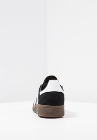 Zapatilla de ante negra con acentos en cuero blanco, tres rayas blancas y suela de goma marr�ón texturizada, vista desde la parte posterior.