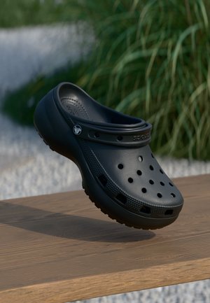Chaussure sabot Crocs noire flottant au-dessus d'une surface en bois avec des plantes vertes et des cailloux blancs en arrière-plan.