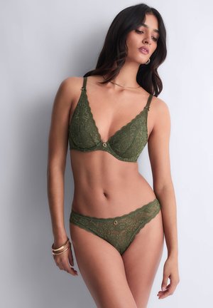 Femme posant dans un ensemble de lingerie en dentelle vert olive avec des détails en forme d'anneau doré, portant des bracelets dorés et des boucles d'oreilles créoles sur un fond clair.
