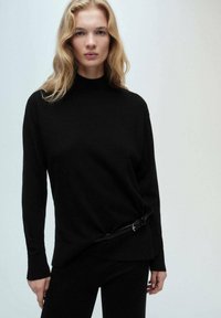 Crna turtleneck veste od mekog pletiva, s detaljem remena u struku, dugim rukavima i opuštenim krojem.