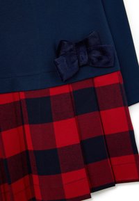 Navyblaues Oberteil mit Samtbogen-Applikation, kombiniert mit einem gestreiften plissierten Rock oder Kleid aus rot- und navyblauem Karomuster.