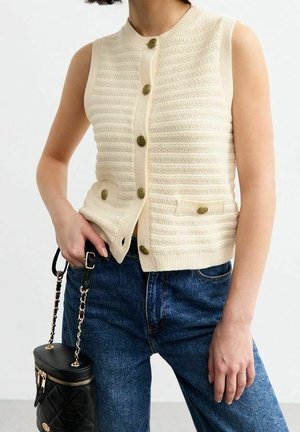 Gilet tricoté crème sans manches avec boutons et poches avant, associé à un jean bleu, tenant un sac à main noir matelassé avec une bandoulière chaîne dorée.
