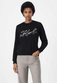 Sudadera negra con cuello redondo y puños de canalé. Presenta el texto bordado en blanco "Karl" con acentos florales, combinada con pantalones gris claro.