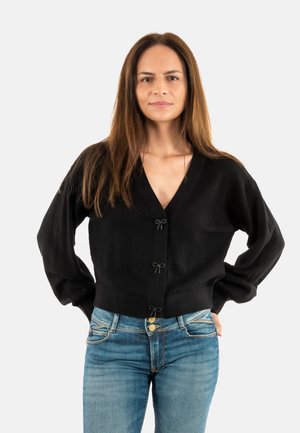 Zwarte gebreide cardigan met een V-hals, voorzien van lange mouwen en decoratieve strikjes bij de sluiting. G gedragen over blauwe spijkerbroeken.