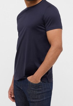 Homme portant un t-shirt bleu marine à manches courtes et col rond, une main dans la poche d’un jean bleu foncé, sur un fond uni.