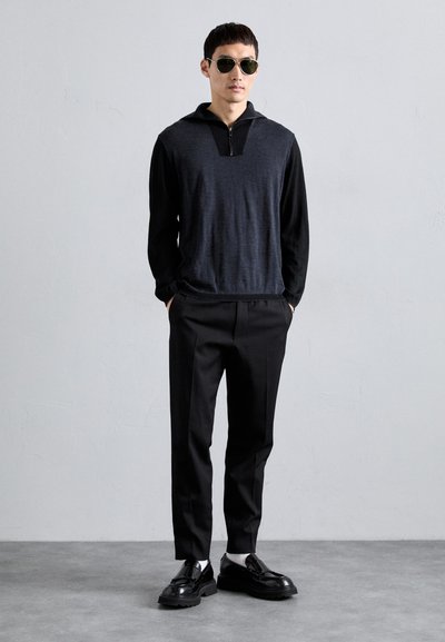 Férfi fekete és szürke cipzáros pulóverben, fekete nadrágban, fekete loafers cipőben fehér zoknikkal, és kerek sötét napszemüvegben áll, kezét a zsebében tartva.