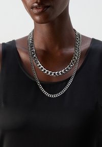 Three layered silver chain necklaces with varying thicknesses, featuring a polished metal finish, worn over a black sleeveless top.