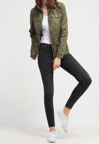 Olivově zelená bunda se čtyřmi předními kapsami, zipem a stojáčkem, spárovaná s černými slim džíny a bílými teniskami.