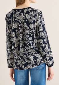 Femme portant un chemisier bleu marine à manches longues avec un imprimé floral blanc et un jean denim bleu, vue de dos sur un fond uni.