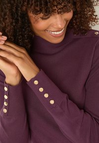 Lila Strickpullover mit hohem Kragen. Verfügt über goldene Knopfdetails an den Bündchen und einen strukturierten Stoff, der den Körper eng umschließt.