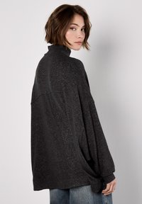 Jeune personne avec les cheveux bruns courts portant un pull ample à col montant gris foncé et un jean bleu, tournée de profil contre un fond léger uni.