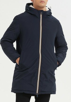 Homme portant un manteau d'hiver à capuche bleu marine avec une fermeture éclair rayée, les mains dans les poches, debout devant un fond clair uni.