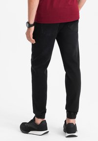 Svarta joggarbyxor med elastiska muddar och två bakfickor, matchade med svarta sneakers med vit sula, bärs med en vinröd skjorta.