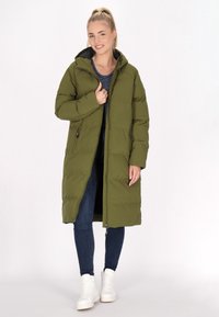 Lång olivgrön pufferjacka med huva, dragkedja framtill och sidofickor; ihopkopplad med mörka jeans och vita högklackade sneakers.