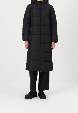 Personne portant un long manteau matelassé noir avec une fermeture éclair à l'avant et une capuche, associé à un pantalon noir à jambes larges et des chaussures noires épaisses.