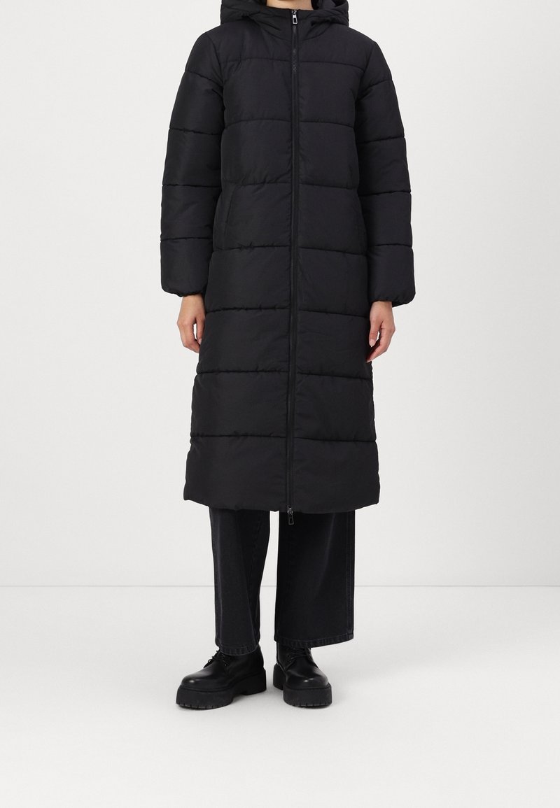 Personne portant un long manteau matelassé noir avec une fermeture éclair à l'avant et une capuche, associé à un pantalon noir à jambes larges et des chaussures noires épaisses.