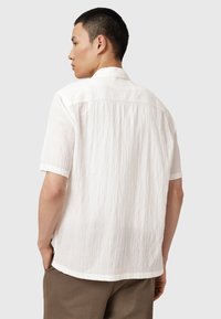 Chemise blanche à manches courtes avec un tissu texturé. Présente un col pointu et un ourlet droit, vue de dos avec des mains dans les poches.