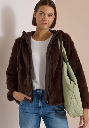 Jeune femme portant une veste marron duveteuse et un jean bleu, tenant un sac fourre-tout matelassé vert clair sur l'épaule, sur un fond uni.