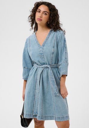 Frau mit lockigem Haar, die ein hellblaues Jeanskleid mit Gürtel trägt und eine schwarze Handtasche hält, steht vor einem schlichten Hintergrund.