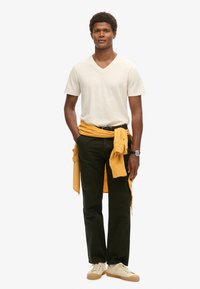 Maglietta crema a V, pantaloni verdi scuri, maglione giallo annodato in vita e sneakers chiare con suola lavorata. Outfit casual con colori neutri.