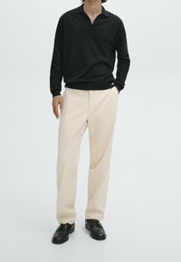 Zwarte polo met lange mouwen en een v-hals, gecombineerd met crèmekleurige tapered broek en zwarte veterschoenen. Zacht materiaal met een ontspannen pasvorm.