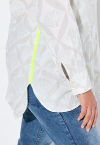 Lichtgekleurde shirt met een schelpprint, voorzien van een heldergele accent langs de zijnaad. Gecombineerd met gestructureerde blauwe jeans.