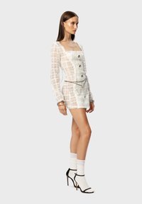 Weißes, strukturiertes Minikleid mit langen Ärmeln, quadratischem Ausschnitt und vorderen Knöpfen. Akzentuiert durch einen silbernen Gürtel. Schwarz-weiße Absatzsandalen.