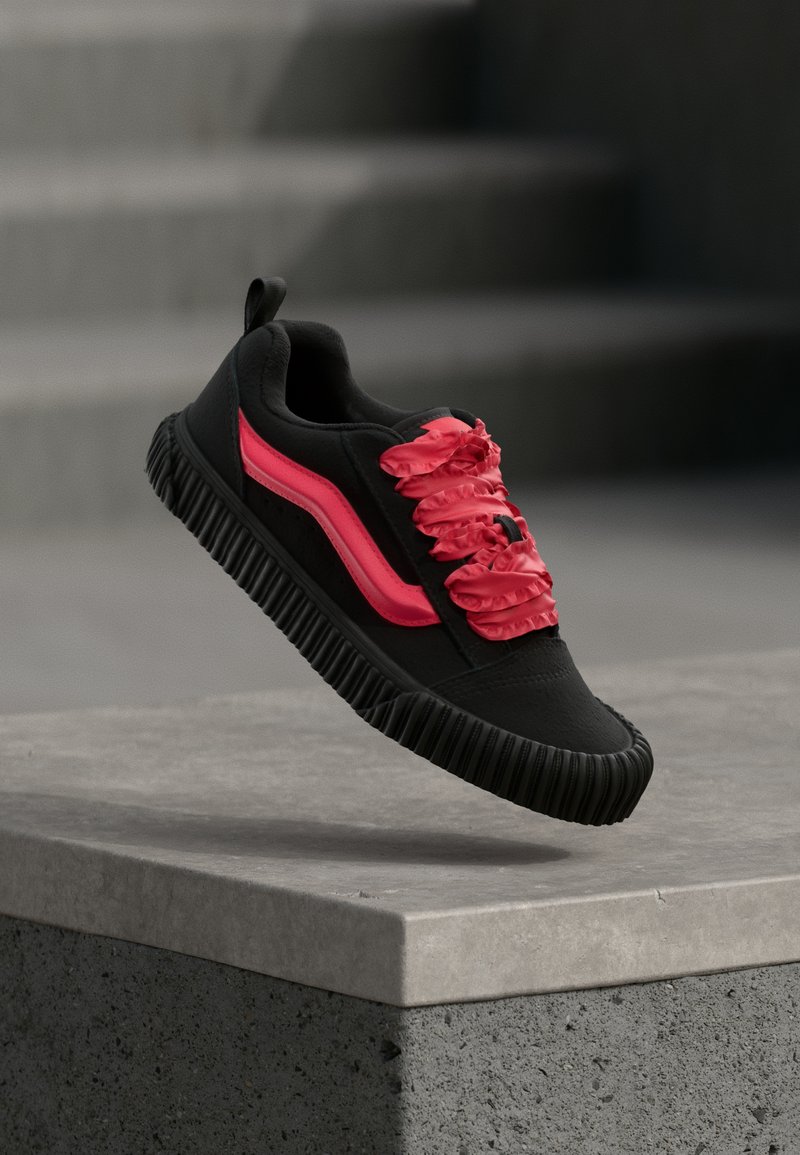 Baskets noires à surface texturée, avec un design de lacets froufroutants roses et une bande rose sur le côté. Semelle en caoutchouc épaisse noire.