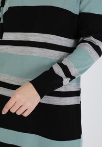 Pull rayé présentant un mélange de couleurs noir, gris et sarcelle, avec des poignets côtelés et une texture tricotée lisse. Main posée sur la manche.