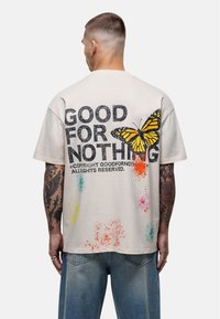 Beżowy t-shirt z krótkim rękawem z dużym graficznym motylem oraz napisem "NIEPRZYDATNY" wytłoczonym na czarno w stylu vintage. Dodatkowe plamy kolorów dodają szczegółów.