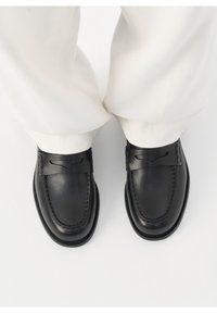 Mocassins penny en cuir noir portés avec un pantalon blanc, vus de dessus sur un fond blanc.