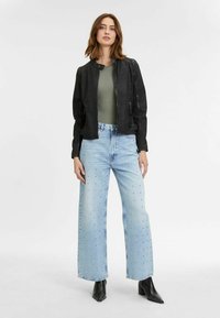 Schwarze Bomberjacke, hellgrüner Body, hellblaue weite Jeans mit metallischen Verzierungen und schwarze Knöchelstiefel. Glatte Texturen.