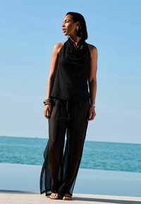 Top nero senza maniche con scollatura a cowl e tessuto testurizzato, abbinato a pantaloni ampi neri trasparenti. Sandali piatti semplici completano il look.