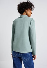 Polo à manches longues en tissu doux et léger vert clair avec un col et une texture lisse, associé à un jean en denim bleu.