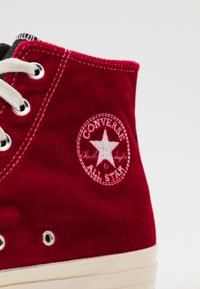 Sneaker alto in velluto rosso con occhielli bianchi, suola in gomma e un patch rotondo con una stella e la scritta "Converse All Star."