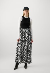 Svart linne över en vit långärmad tröja, kombinerat med en maxikjol med svartvit mönstring och svarta ankelboots. Innehåller en svart keps.