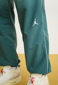Teal trainingsbroeken met witte piping en een wit Jumpman-logo, gedragen met witte en rode Air Jordan sneakers op een mosterdkleurige vloer.