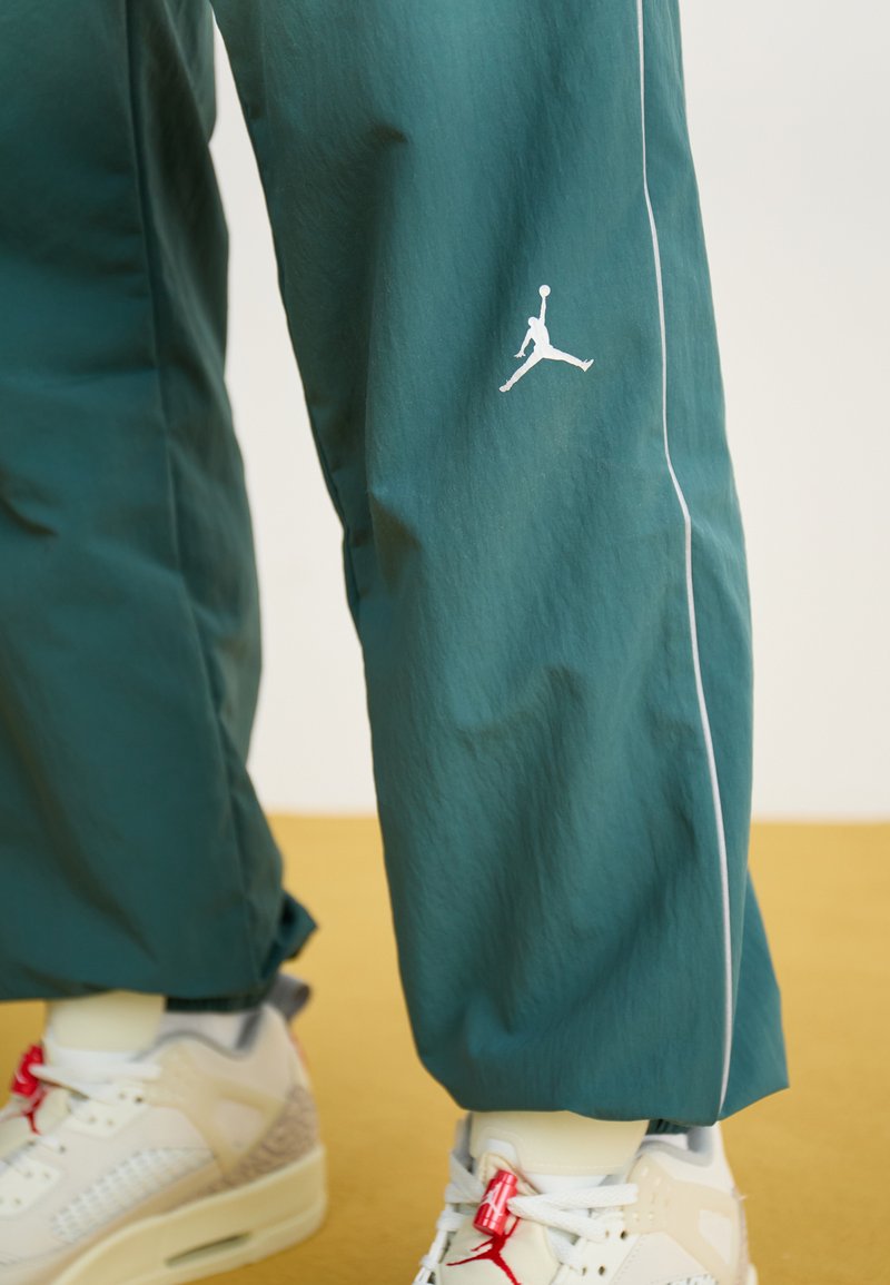 Turkusowe dresy z białymi wstawkami i białym logo Jumpman, noszone z białymi i czerwonymi butami Air Jordan na musztardowej podłodze.