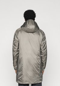 Hosszú, könnyű szürke parká, kapucnival. Simább, fényes szövet, amely diszkrét textúrával rendelkezik. Az ujjak a mandzsetta résznél enyhén összegyűrtek.